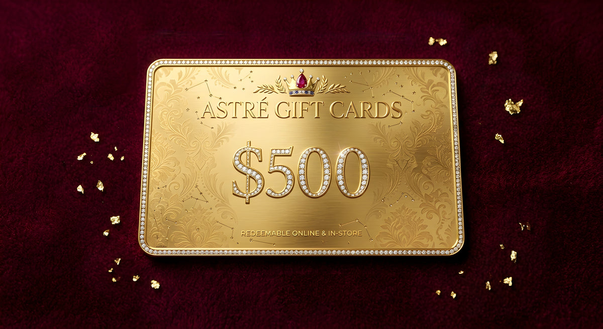 A rectangular gold gift card labeled “ASTRÉ GIFT CARDS” and “$500” with decorative embossed patterns and a small crown emblem, placed on a dark red textured background with scattered gold leaf pieces.